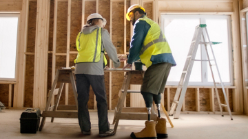 Un travailleur de la construction ayant un handicap physique discute de plans avec une collègue autour d’une table de travail à l’intérieur d’une maison en construction.