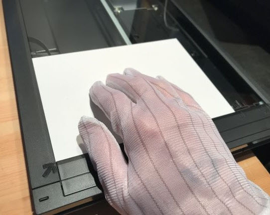 Image of a gloved hand placing an IT8 target face down on the glass platen. Only the white backing of the target is visible.