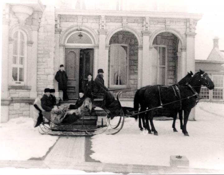 People sitting in sleigh pulled by two horses in front of house