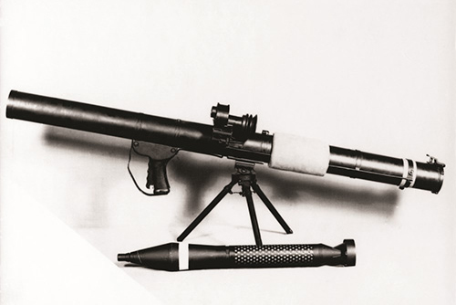 Photo en noir et blanc d’une arme antichar montée sur trépied, munitions à ses côtés, sur fond blanc.