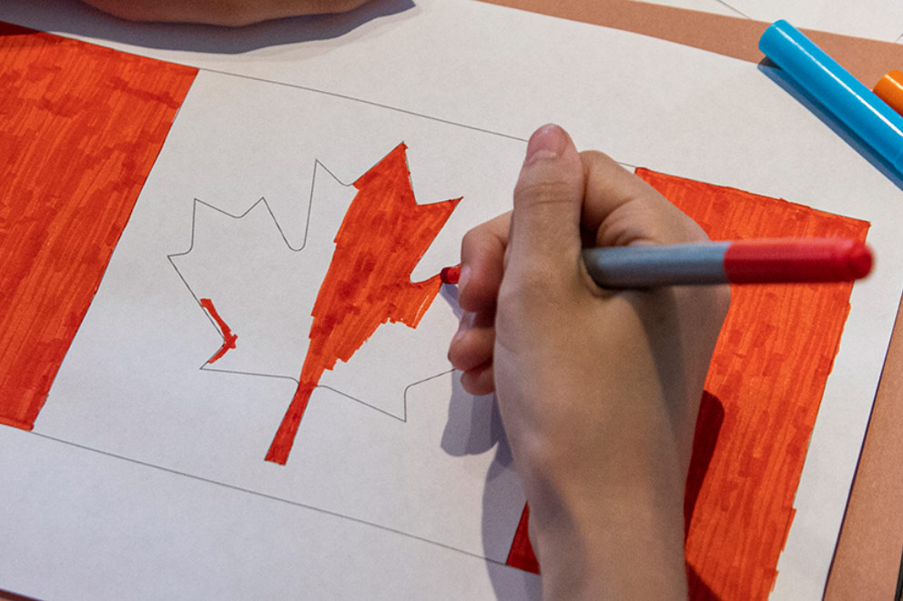 Close up of a hand holding a red marker coloring the inside of the Canadian Flag maple leaf drawn on a white piece of paper.