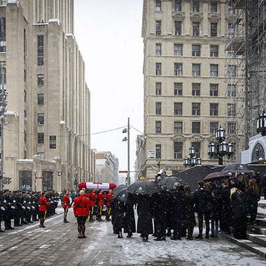 A small crowd of people and uniformed officers in formation stand as a casket is carried by uniformed pallbearers.

