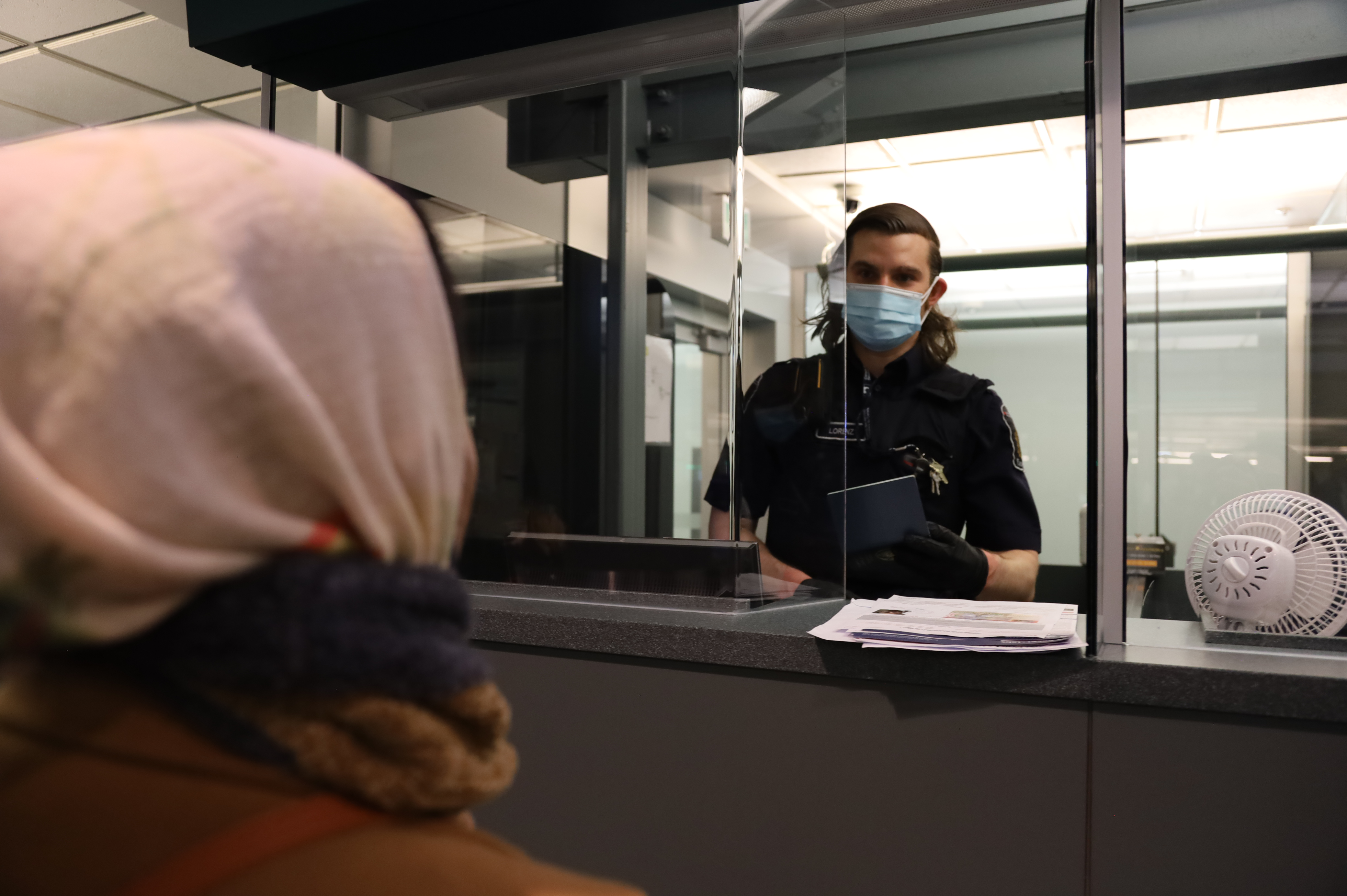 Inside a booth, a border services officer wearing a mask speaks to an individual while inspecting their passport