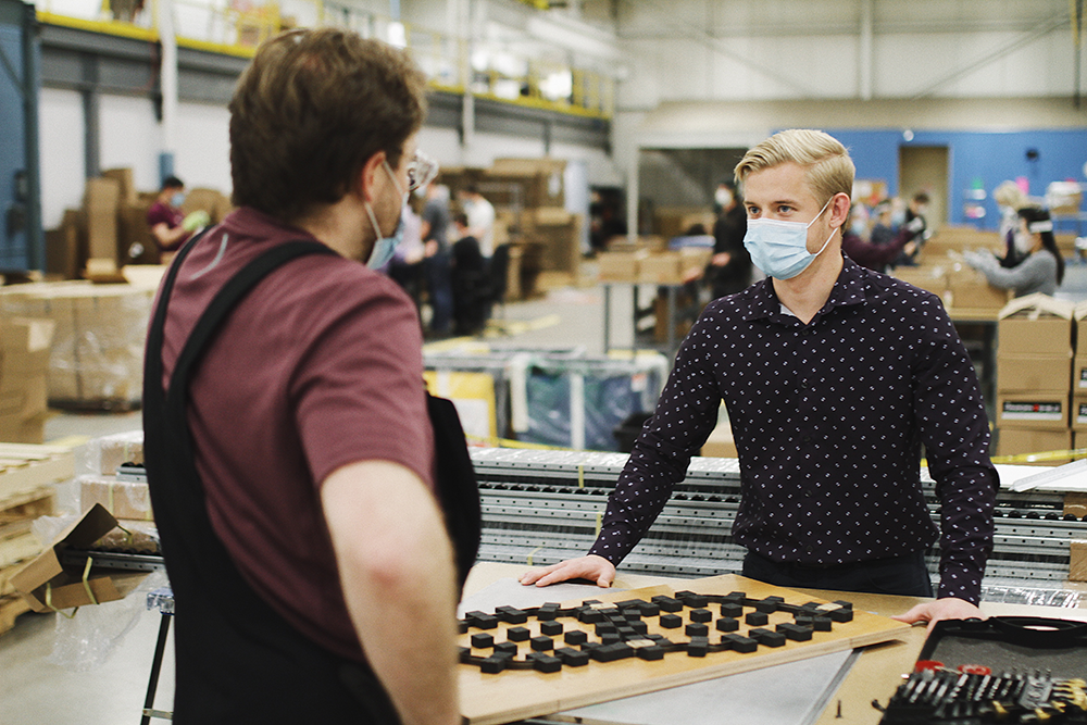 Jeremy Hedges and a person, are wearing masks and facing each other. On a table between them are tools and a plank of wood. Behind them, there are several people working in a factory setting.