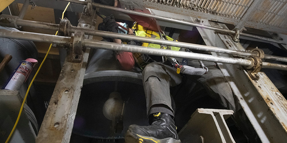 Vue en contre-plongée d'une personne travaillant sur une cloche massive dans un environnement confiné et entouré de tuyaux.